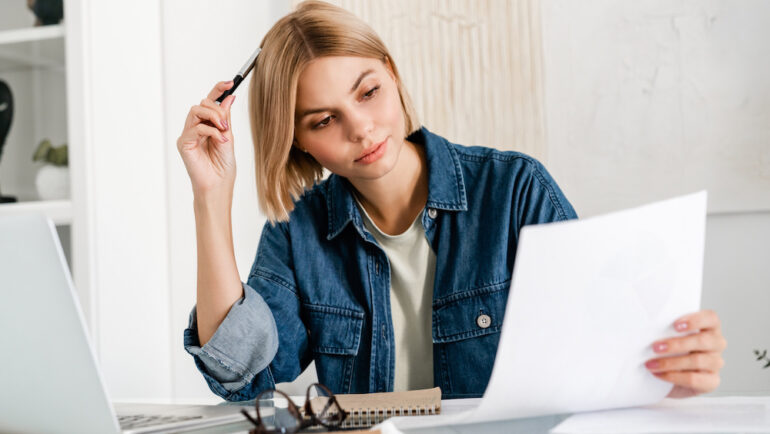 The pensive renter is reviewing a new lease showing a rent increase. and working on the laptop. Caucasian young woman holding a document dealing with the problem.